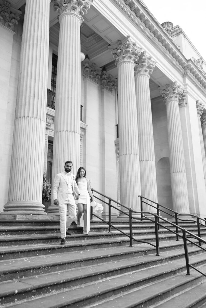 A couple dressed in formal wedding attire walks down the steps of a grand building with tall columns and ornate architecture. The woman holds a bouquet and they appear happy and relaxed. The image is in black and white.