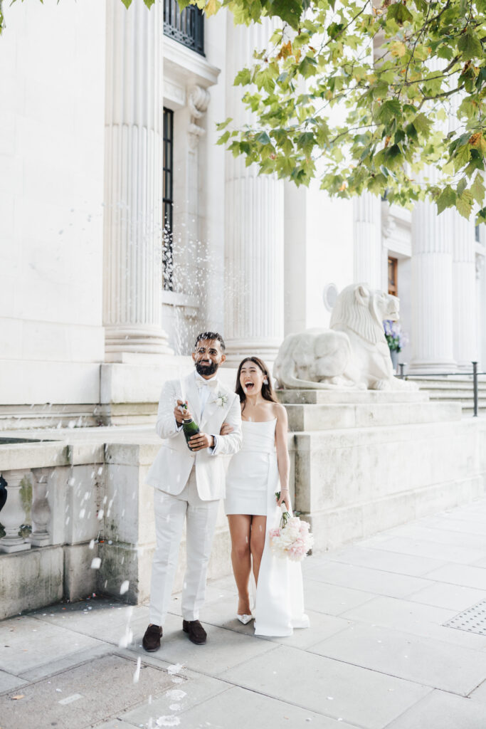 A couple dressed in white stands outside a grand building, smiling and celebrating as the man pops a bottle of champagne, causing it to spray. The woman holds a bouquet and laughs under leafy tree branches.