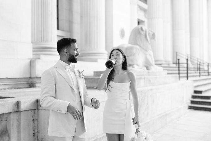 A man in a light suit and a woman in a white dress stand outside a building with columns; the woman is drinking from a bottle and holding a bouquet, while the man smiles at her.