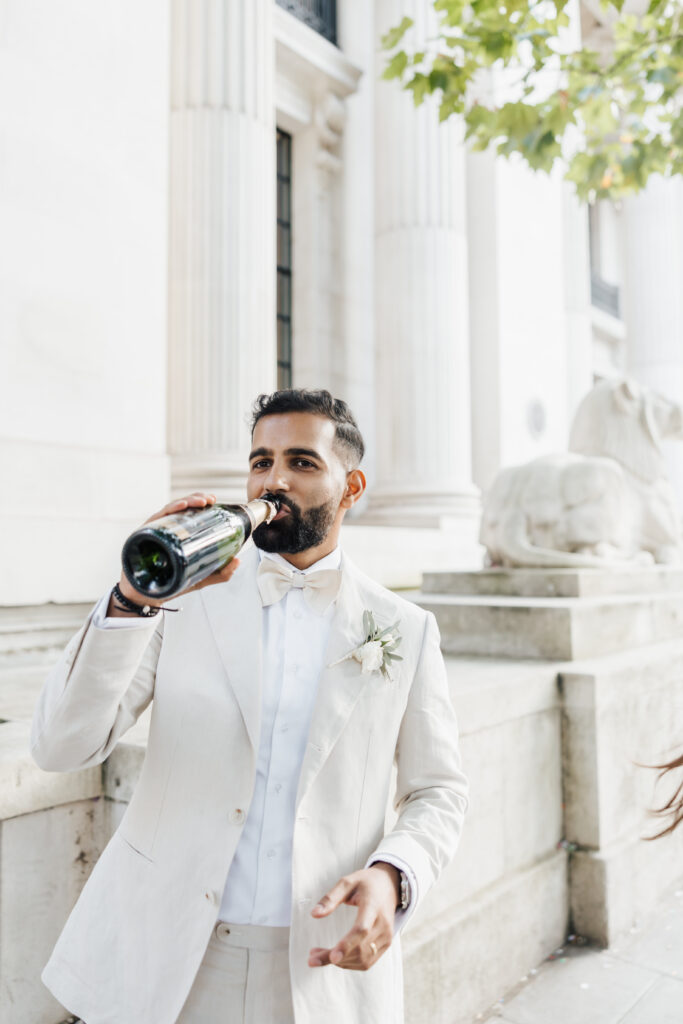 A man in a light suit and bow tie, with a boutonnière on his lapel, drinks from a champagne bottle outside a white stone building with columns and a lion statue.
