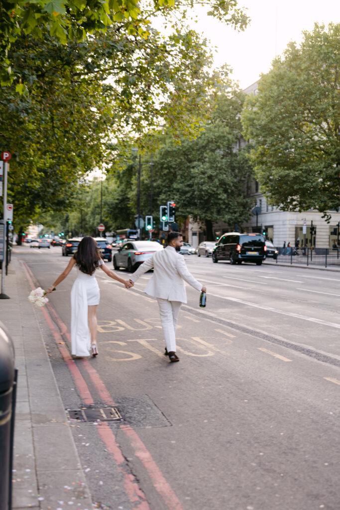A couple dressed in white, holding hands and walking down a city street. The woman holds a bouquet, and the man has a bottle. Cars are visible on the road and trees line the street.