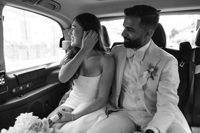 A bride and groom sit together in the backseat of a car. The bride is smiling and adjusting her hair, while the groom looks at her with a smile. Both wear formal wedding attire. The image is in black and white.