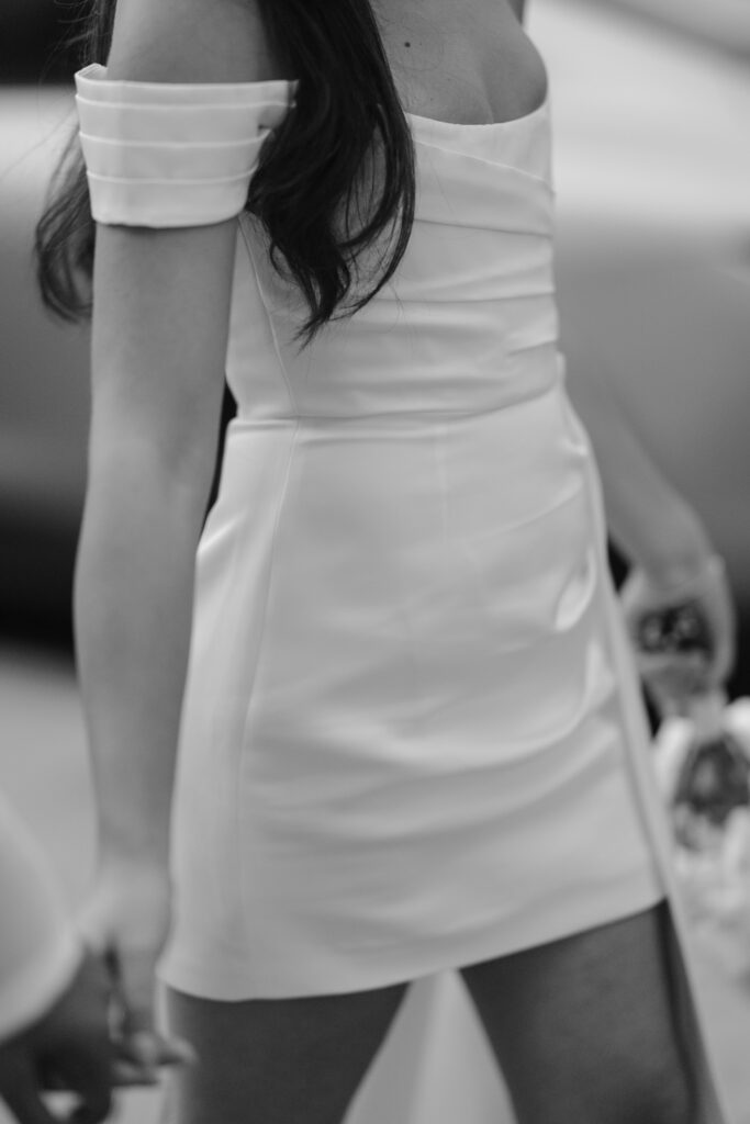 A woman in an off-the-shoulder, fitted, white dress holds someones hand and a small clear bottle. The photo is in black and white and captures her from the shoulders to mid-thigh.