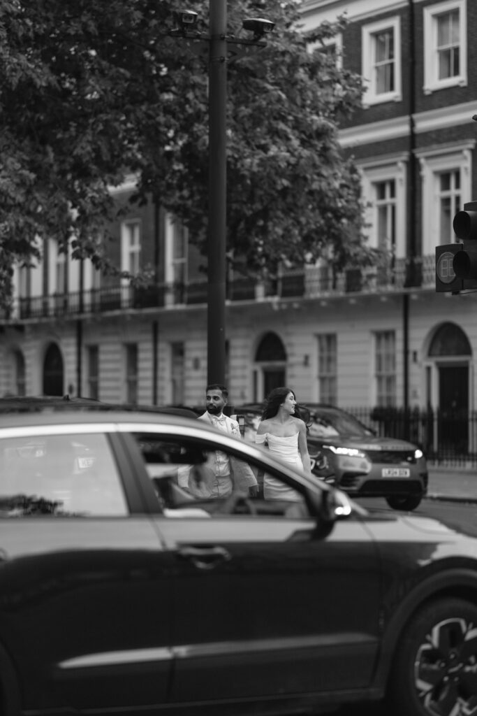 A man and woman cross a city street while cars pass by in the foreground; the image is black and white, with tall buildings and leafy trees in the background.