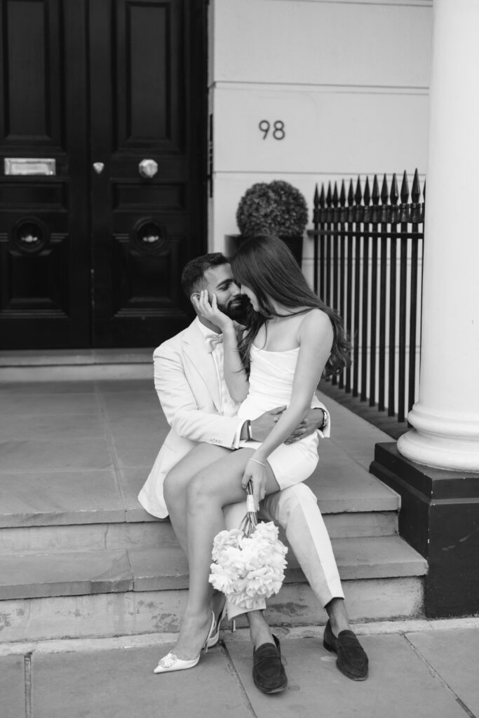 A couple dressed in white sits on steps in front of a door numbered 98. The woman holds a bouquet and sits on the man’s lap as they gaze at each other affectionately. The image is in black and white.