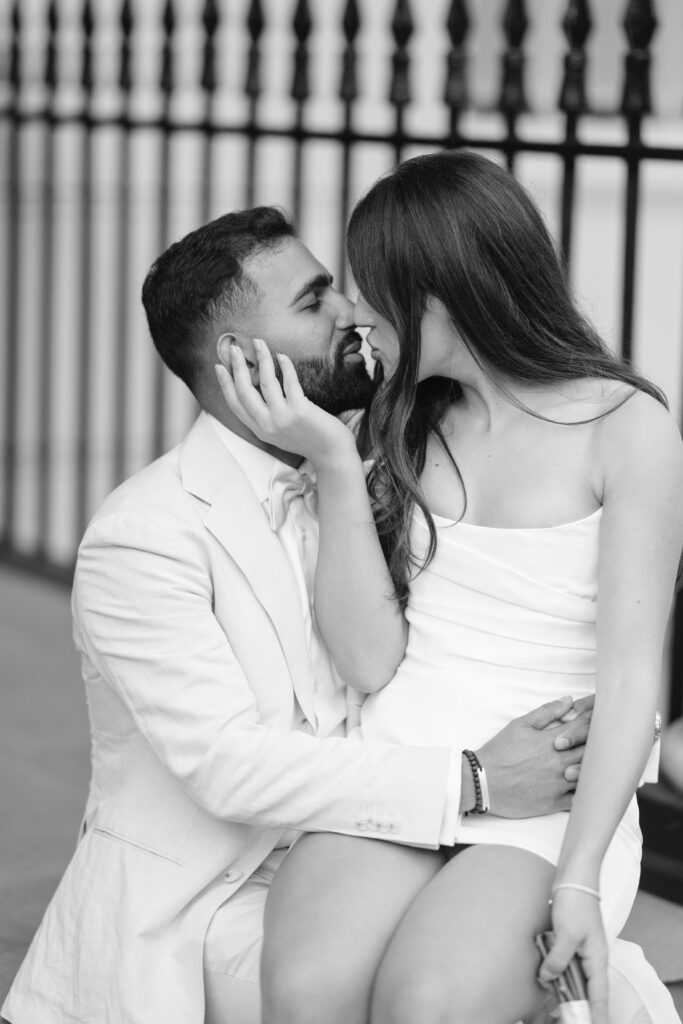 A man in a light suit sits with a woman in a strapless white dress on his lap; they embrace closely and almost kiss, gazing into each others eyes. The photo is black and white with a blurred fence in the background.
