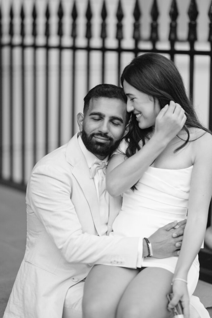 A couple sits closely together, both dressed in white. The man has his eyes closed and smiles softly as the woman laughs, gently touching her hair. A black iron fence is visible in the blurred background.