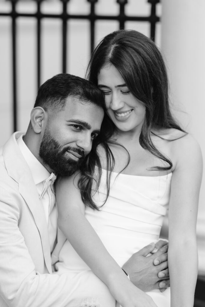 A couple sits closely together, the man leaning his head on the womans shoulder while she smiles with her eyes closed. Both are dressed formally in light-colored outfits, and appear happy and relaxed. The image is in black and white.