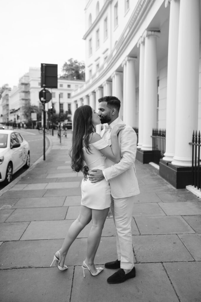 A couple dressed elegantly, the woman in a short dress and heels and the man in a light suit, share an intimate moment on a city sidewalk in front of a white building with columns. The photo is black and white.