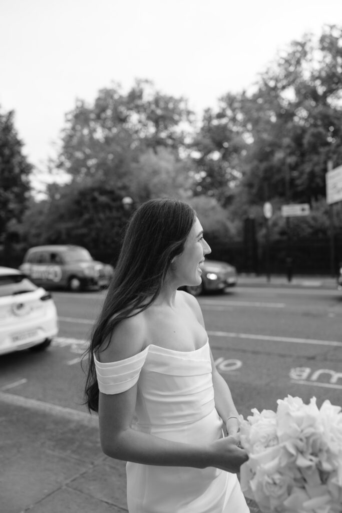 A woman in an off-the-shoulder white dress stands outdoors, smiling and holding a bouquet. Cars and trees line the street in the background. The photo is in black and white.