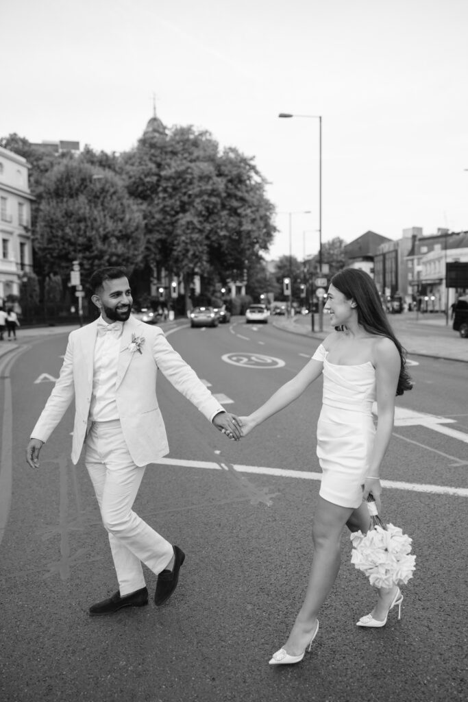 A couple dressed in wedding attire holds hands and walks across a city street. The man wears a light-colored suit, and the woman wears a short white dress and holds a bouquet. Cars and trees are visible in the background.