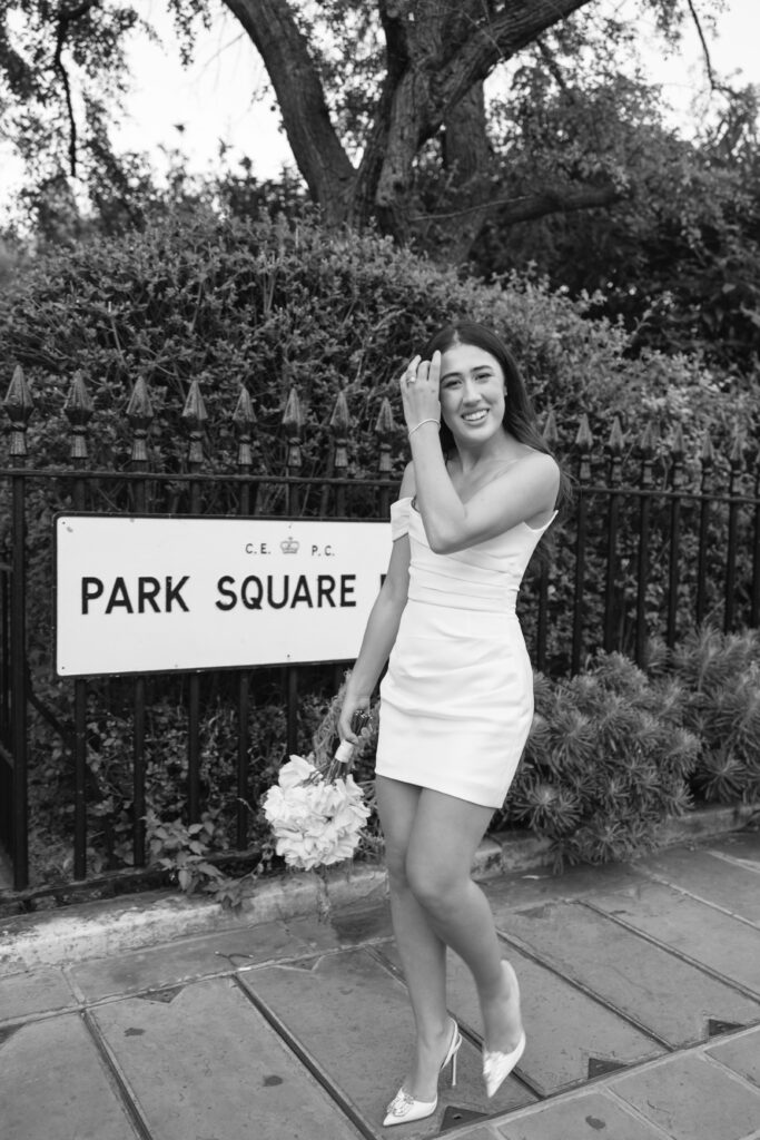A woman in a short, off-the-shoulder dress and heels stands on a sidewalk by a Park Square sign, holding a bouquet and smiling with one hand in her hair; trees and a fence are in the background.