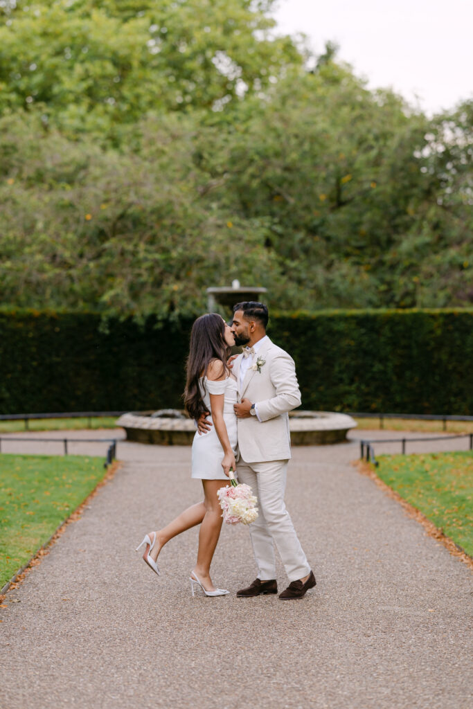 A couple dressed in light-colored outfits embrace and kiss in a garden pathway. The woman is holding a bouquet and standing on one leg, while the man wears a suit. Green trees and a fountain appear in the background.