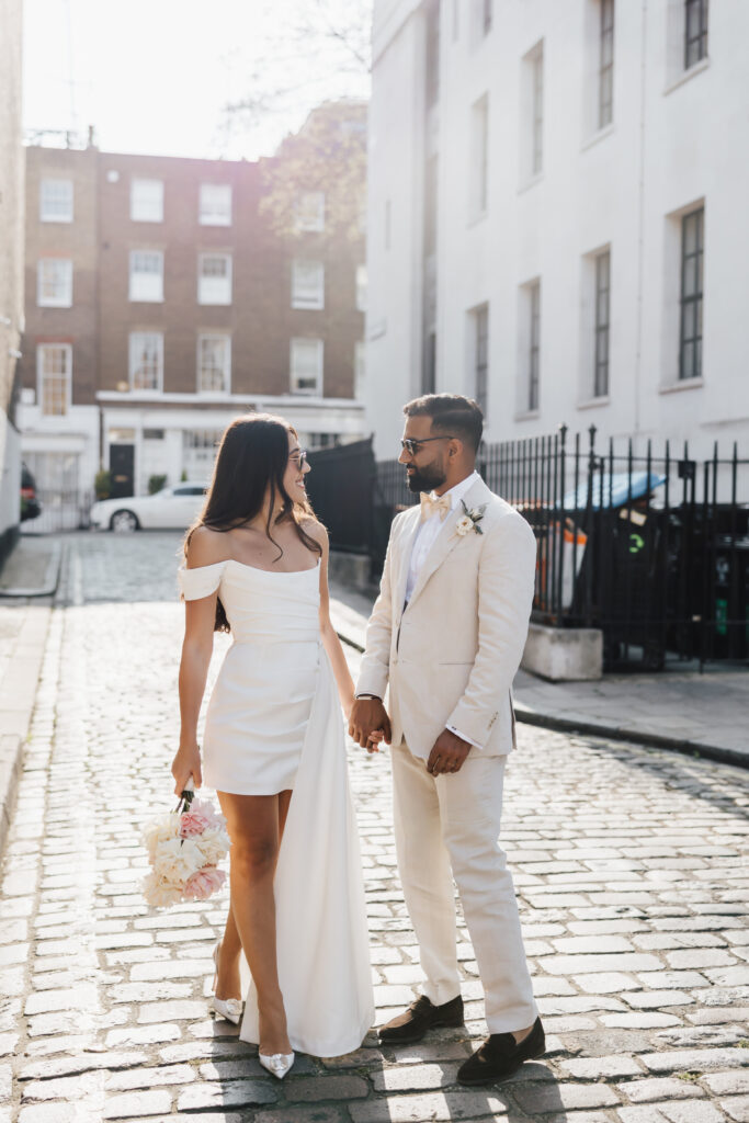 A bride and groom stand on a sunlit cobblestone street, holding hands and looking at each other. The bride wears an off-shoulder white dress and holds a bouquet, while the groom wears a cream suit with a boutonniere.