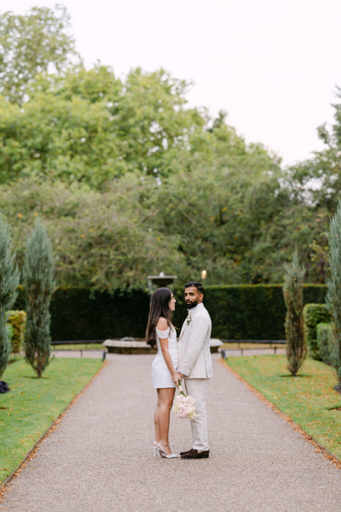A couple stands facing each other on a garden path, holding hands. The woman wears a white dress and heels, and the man wears a light suit. Trees and greenery surround them, and a bouquet is in the woman’s hand.