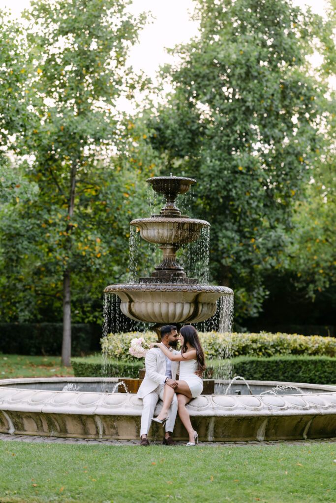 A couple dressed in white sits closely together on the edge of a large tiered fountain in a lush green park, surrounded by trees and hedges, sharing an intimate moment.