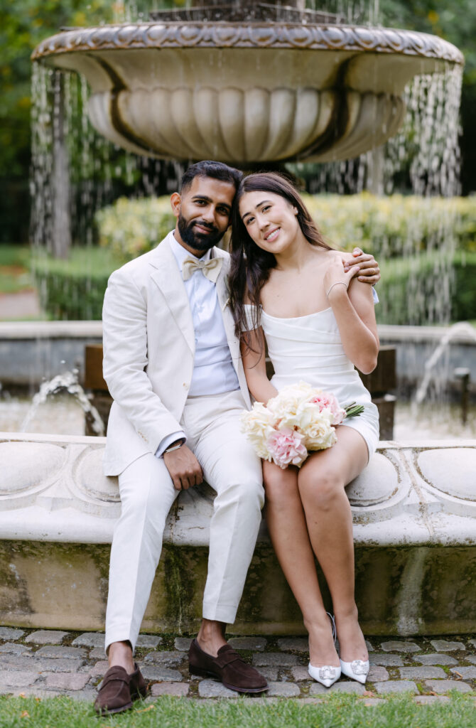 A couple dressed in white sits closely on a stone bench in front of a fountain. The woman holds a bouquet of flowers and leans on the man, both smiling gently at the camera.