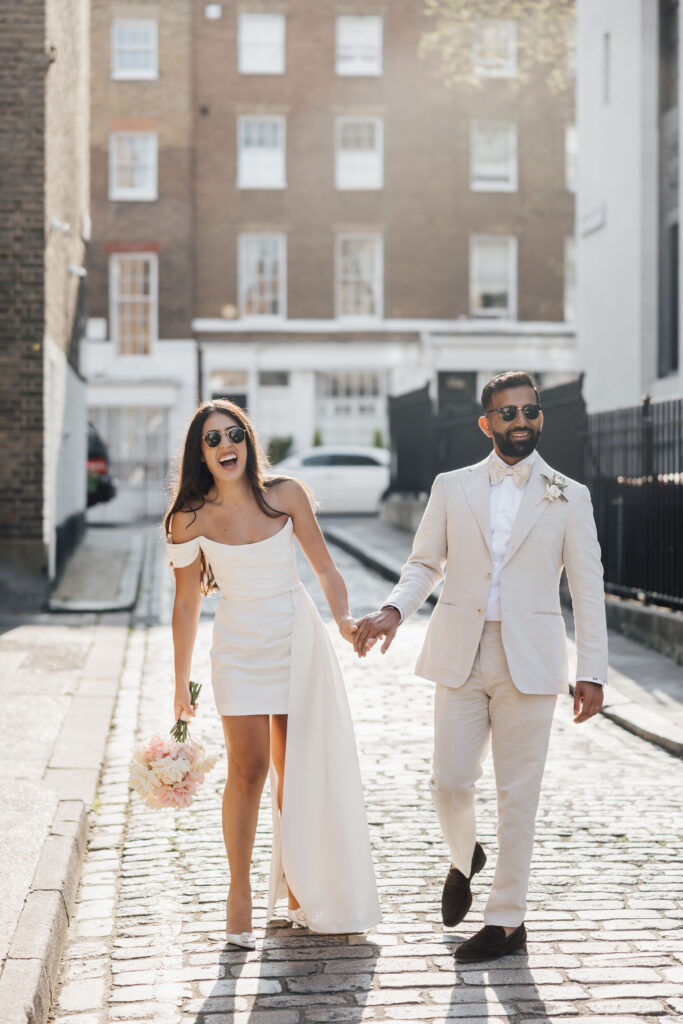 A joyful bride and groom, both wearing sunglasses and dressed in light-colored attire, walk hand in hand on a cobblestone street; the bride holds a bouquet of flowers and smiles brightly.