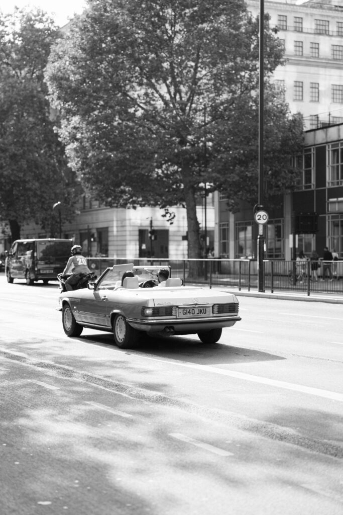 A black and white photo of a vintage convertible car driving down a city street, with a person wearing a helmet riding a motorcycle nearby. Trees and buildings line the background.