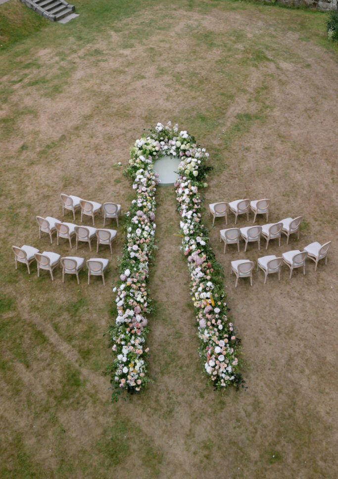 An outdoor wedding ceremony setup features two rows of chairs facing a circular floral altar, with lush flower arrangements forming an aisle and surrounding the altar on a patchy grassy area.