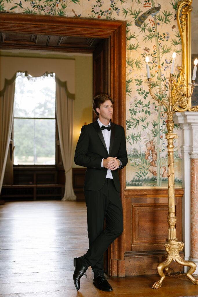 A man in a black tuxedo with a bow tie leans casually against a doorway in an elegant, ornately decorated room with floral wallpaper, a tall gold candelabra, and large windows in the background.