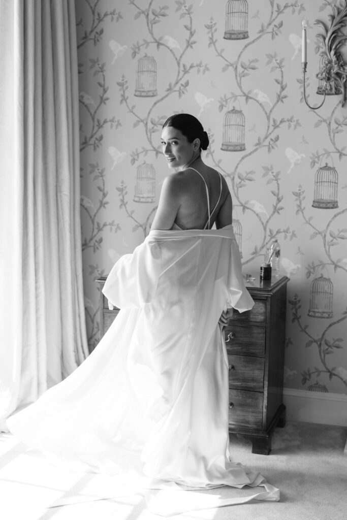 A woman in a long, elegant, backless gown stands by a window and wooden nightstand, gazing over her shoulder. Soft light streams in, and birdcage-patterned wallpaper decorates the room. Black and white photo.