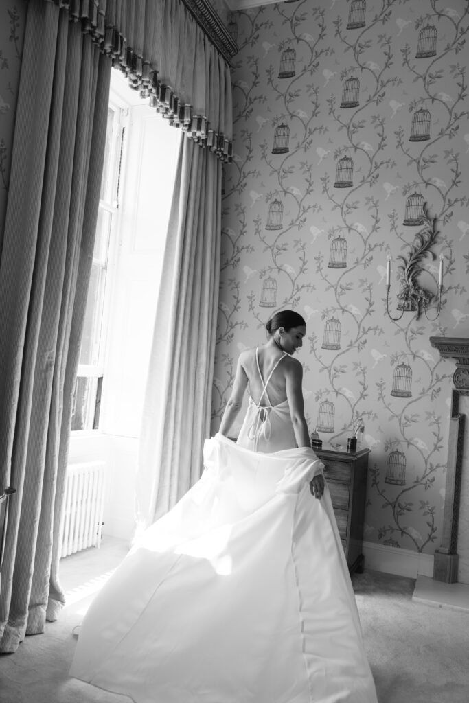 A woman in a backless wedding dress stands by a sunlit window, holding her gown. The room features patterned wallpaper with birdcages, heavy curtains, and elegant furnishings. The photo is in black and white.