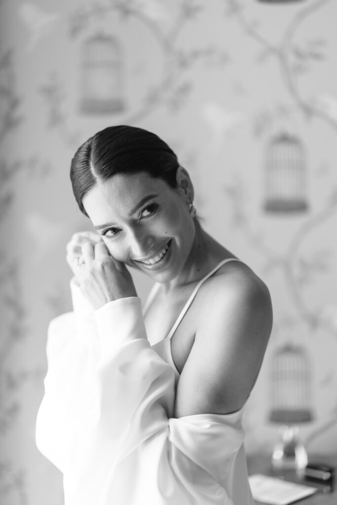 A smiling woman in a white dress adjusts her earring, with her dark hair pulled back. The background features a wallpaper with birdcage designs. The image is in black and white.