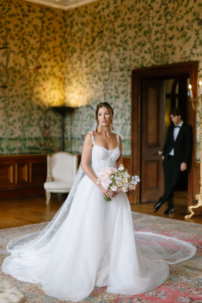 A bride in a white wedding dress holds a bouquet of flowers, standing in an elegant room with floral wallpaper. A groom in a black tuxedo stands in the background near a doorway.