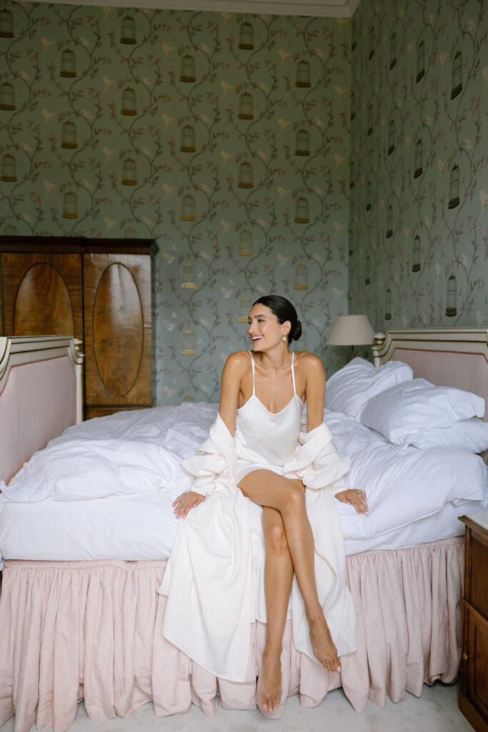 A woman in a white slip dress and robe sits smiling on the edge of a neatly made bed in a vintage-style bedroom with green patterned wallpaper and wooden furniture.