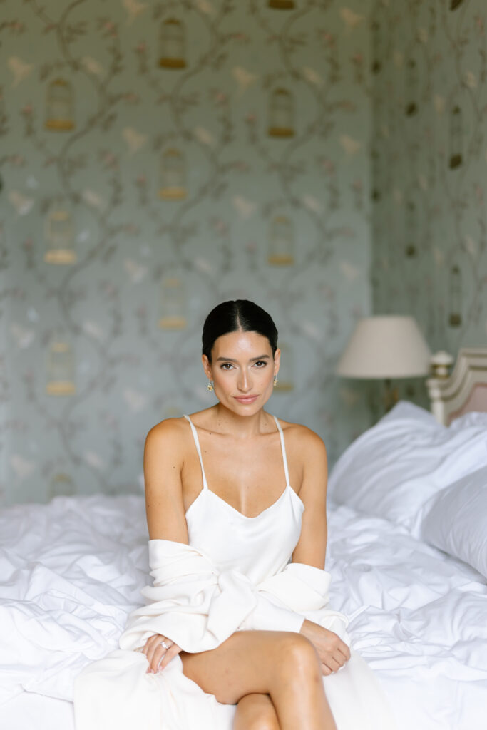 A woman with dark hair in a low bun sits on a bed with white bedding, wearing a white camisole dress and a draped white robe, against a soft green floral wallpaper background. She is looking calmly at the camera.