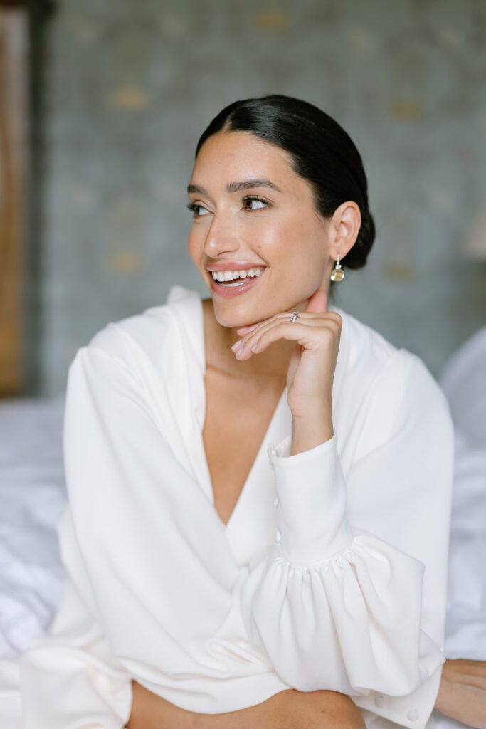 A woman with dark hair styled in a low bun, wearing a white blouse and gold earrings, smiles while resting her chin on her hand. She is seated indoors with a soft-focus background.