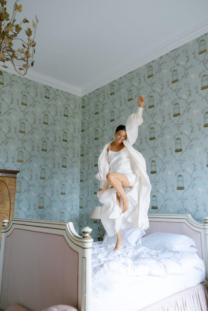 A woman in a white robe and dress joyfully jumps on a bed in a vintage-style bedroom with pale green wallpaper featuring birdcages and branches. The room has elegant, light-colored furniture and a chandelier.