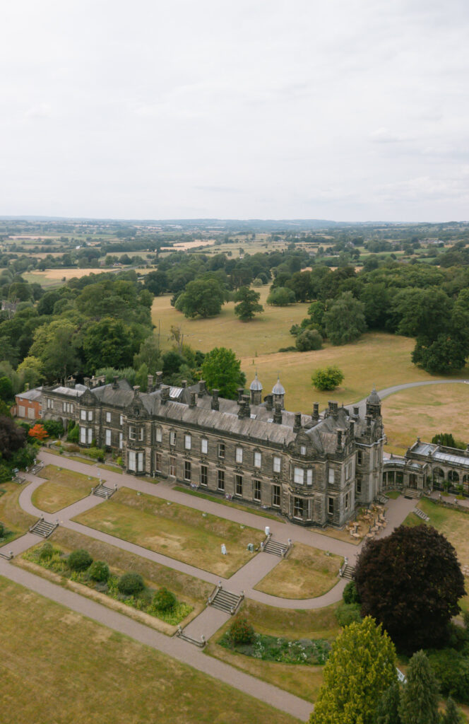 A large historic stone mansion surrounded by landscaped gardens and pathways, set in a vast green countryside with trees, fields, and distant hills under a cloudy sky.
