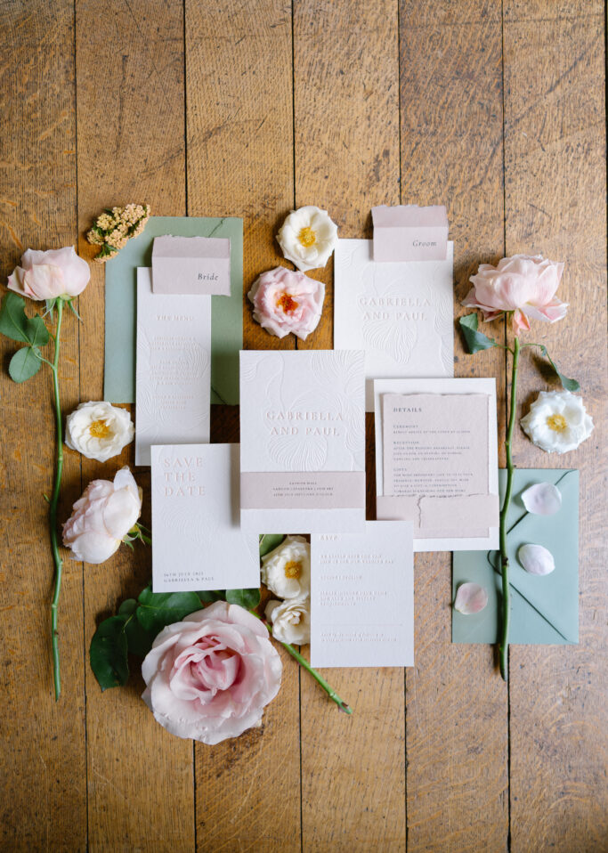 Wedding invitation suite arranged on a wooden floor, surrounded by blush pink and white flowers and green leaves. The set includes various cards and envelopes in soft pastel shades.