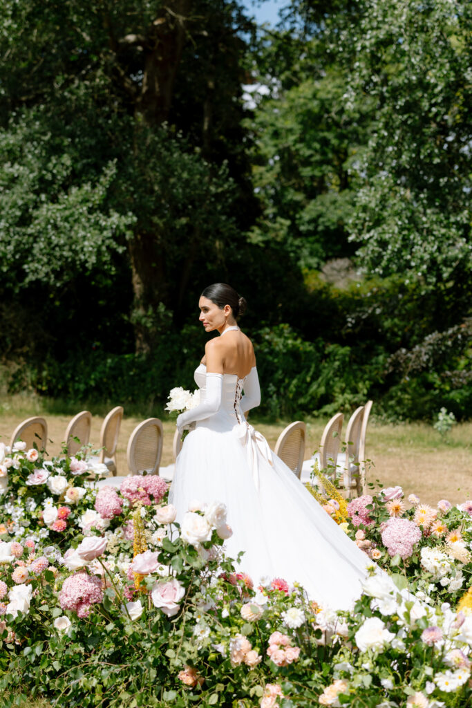 A bride in a white off-the-shoulder gown stands outdoors, holding a bouquet and surrounded by lush pink, white, and peach flowers, with empty chairs and greenery in the background.