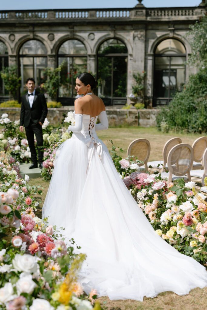 A bride in a white gown with a long train stands among flower arrangements outdoors, looking over her shoulder. An elegantly dressed groom in a tuxedo stands in the background near a historic stone building.