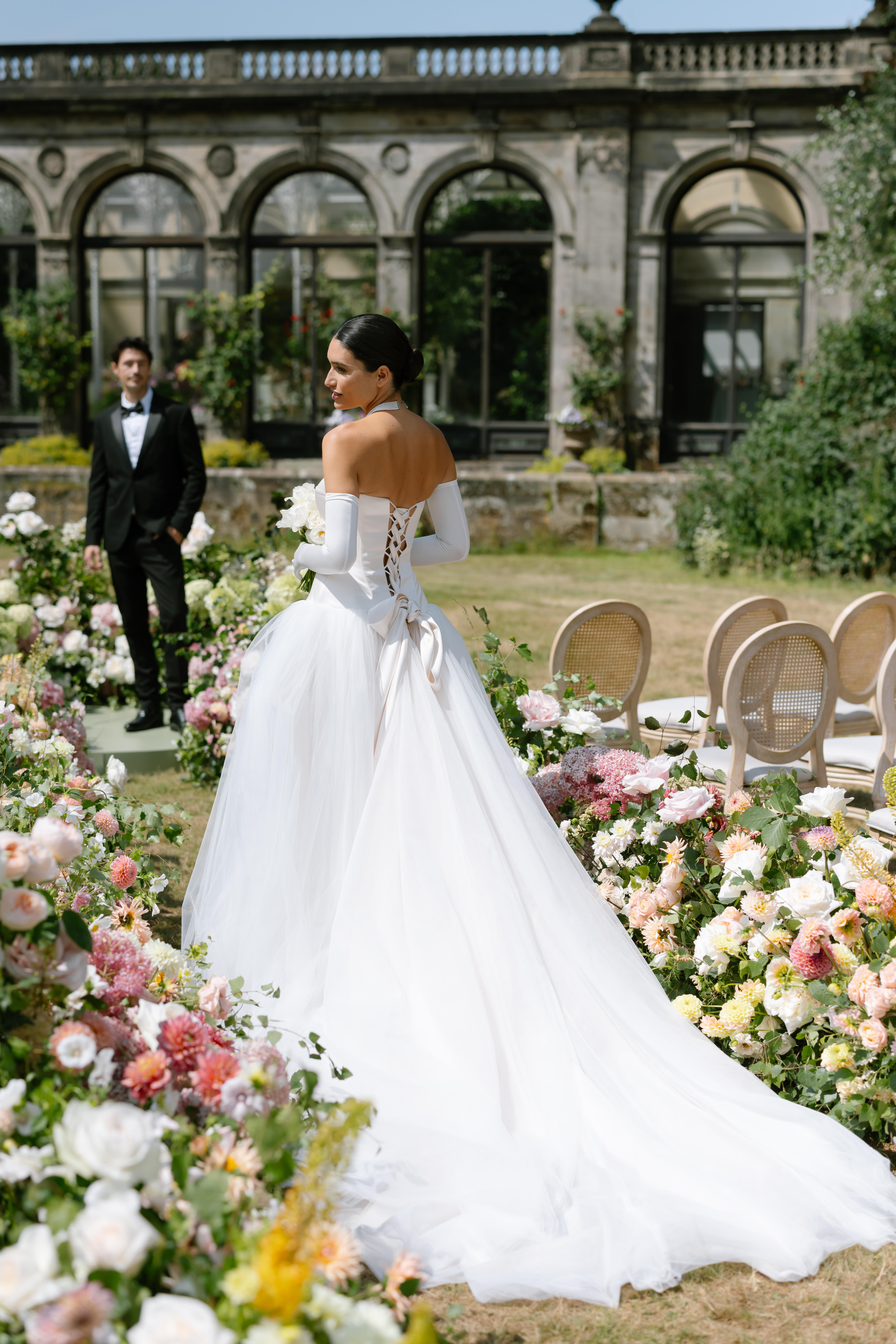 A bride in a white gown with a long train stands among flower arrangements outdoors, looking over her shoulder. An elegantly dressed groom in a tuxedo stands in the background near a historic stone building.