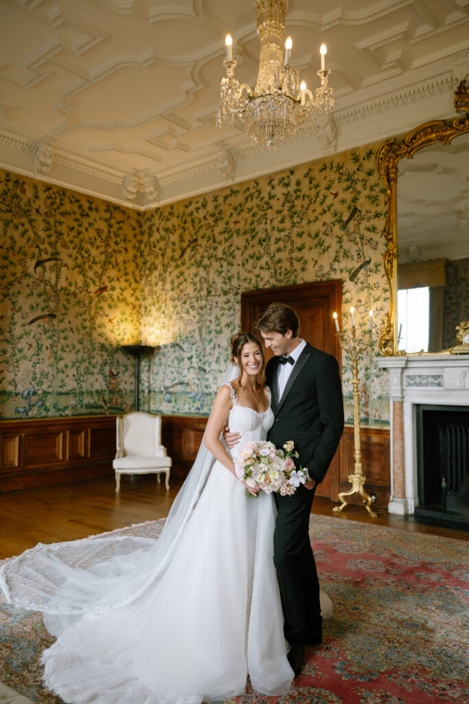 A bride in a white gown and a groom in a black tuxedo stand smiling together in an elegant room with ornate wallpaper, a chandelier, and a large mirror above a fireplace. The bride holds a bouquet of flowers.