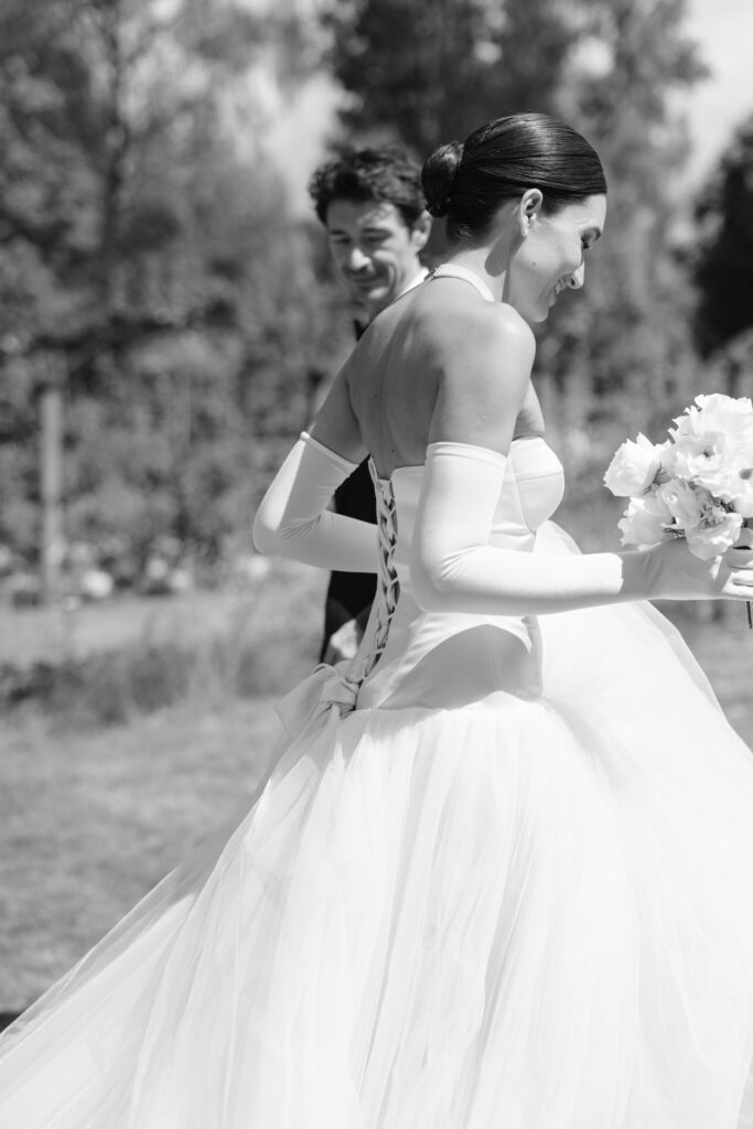 A bride in a strapless gown and long gloves holds a bouquet of flowers and smiles, walking outdoors with a man in a suit beside her. The scene is bright and set in a natural, airy environment.