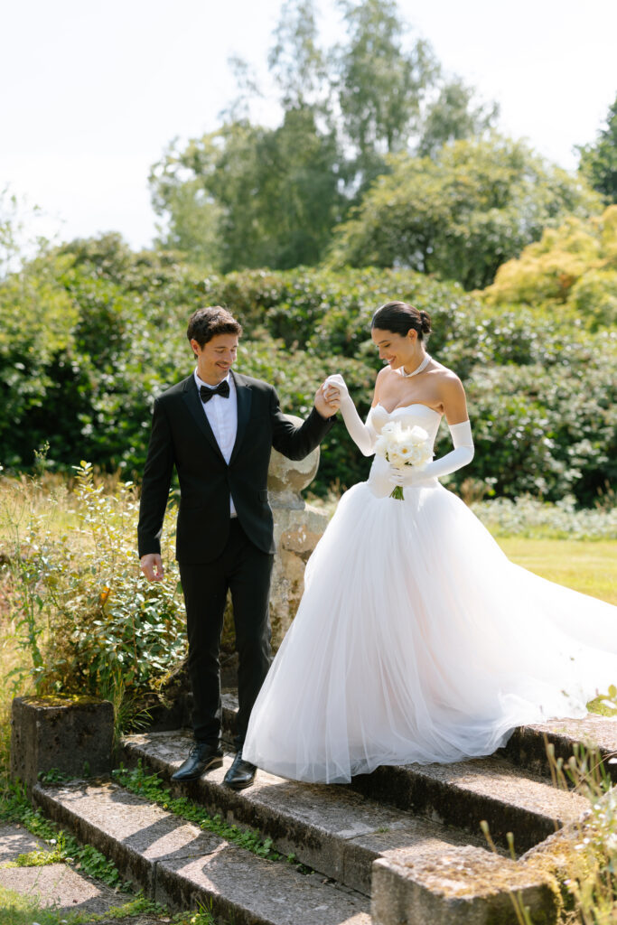A bride in a white gown and gloves holds a bouquet and the hand of a groom in a black suit as they walk down outdoor stone steps, surrounded by greenery and trees on a sunny day.