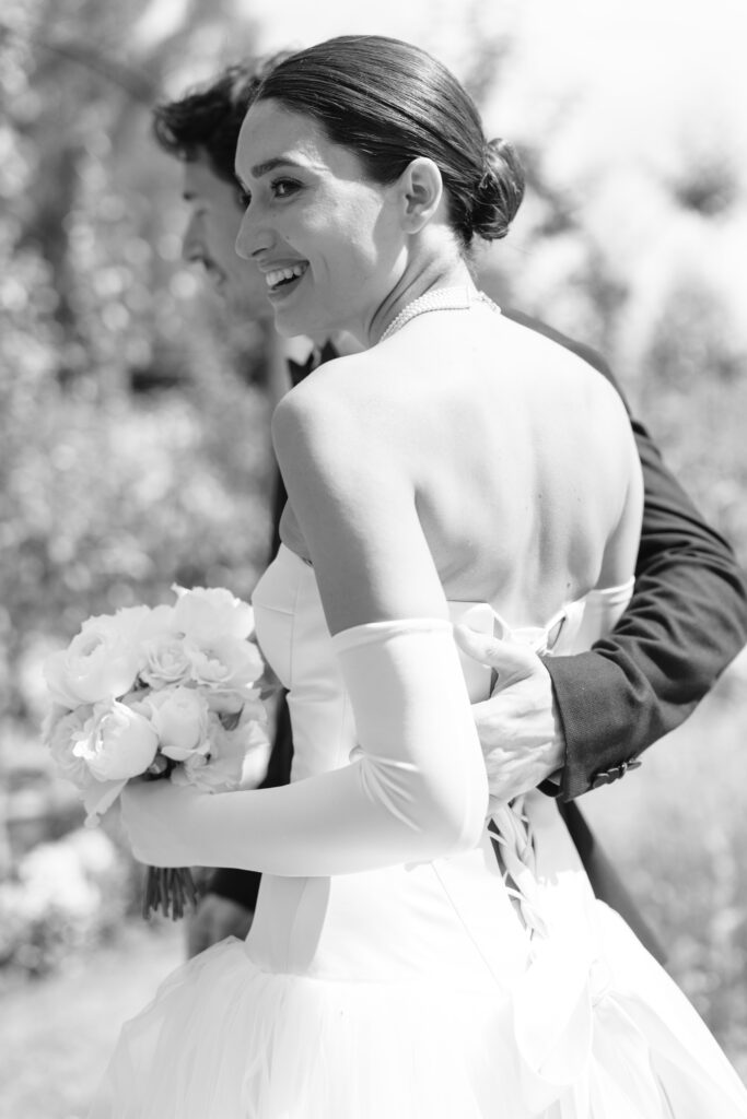 A smiling bride in a strapless wedding gown and long gloves holds a bouquet of flowers while being embraced by a suited person outdoors at their Sandon Hall Wedding. The black and white photo captures pure joy and elegance.