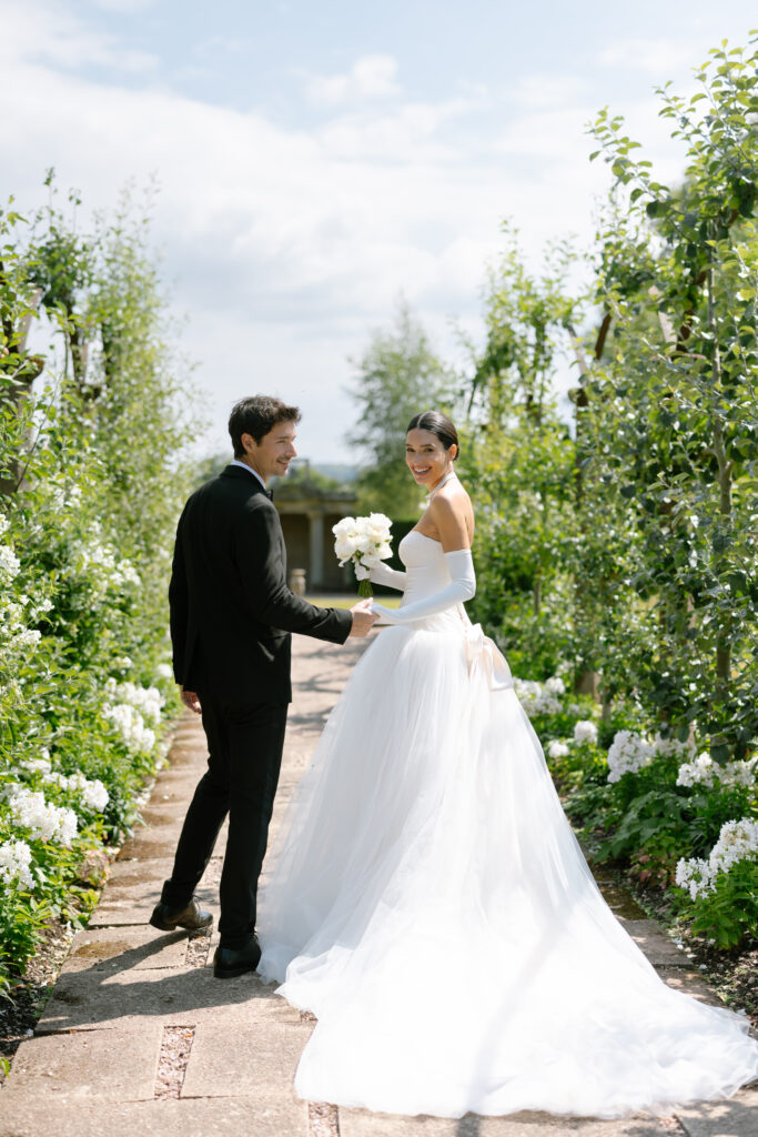A bride in a white gown and a groom in a black suit hold hands and smile at the camera while walking outdoors on a garden path, captured by a Sandon Hall Wedding photographer, surrounded by lush greenery and white flowers.