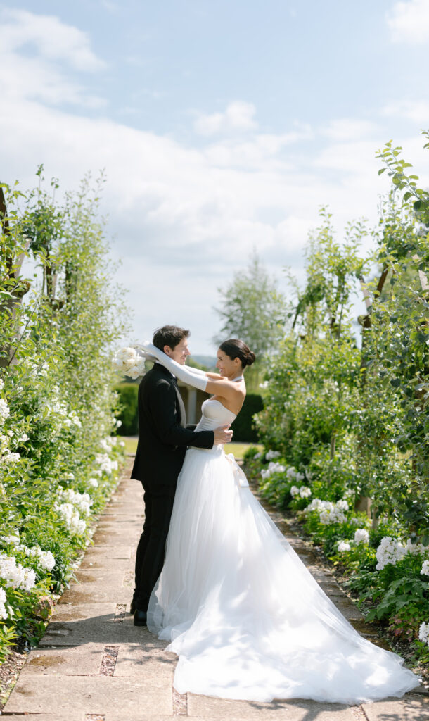 A bride and groom stand facing each other on a garden path lined with green plants and white flowers at their Sandon Hall Wedding, smiling and embracing on a sunny day with a blue sky.
