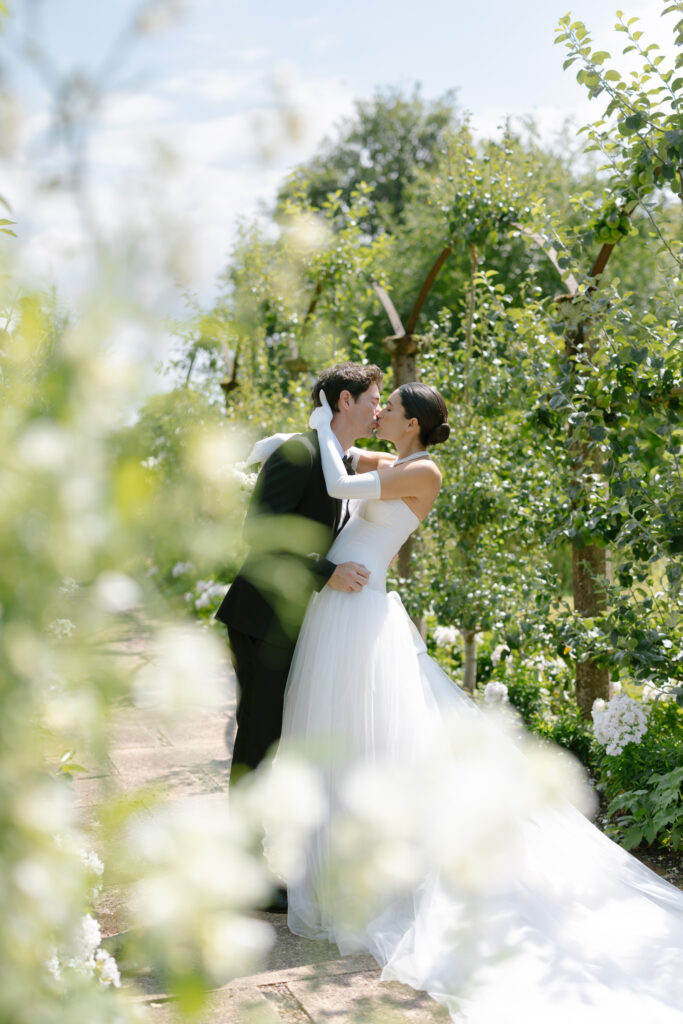 A bride and groom share a kiss outdoors in a lush, green garden, surrounded by blooming flowers and trees. The bride wears a flowing white gown and gloves; the groom is dressed in a dark suit. Sunlight filters through the leaves.