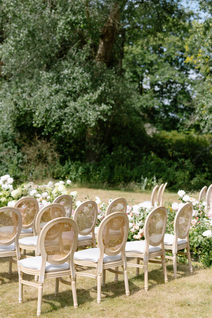 Rows of elegant wooden chairs with white cushions are arranged outdoors on grass, facing a floral arrangement of white and blush flowers, with lush green trees and foliage in the background.