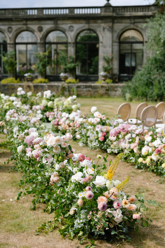 Rows of pastel and white flowers with greenery decorate an outdoor garden setting, with elegant chairs and a stone building with large arched windows in the background.