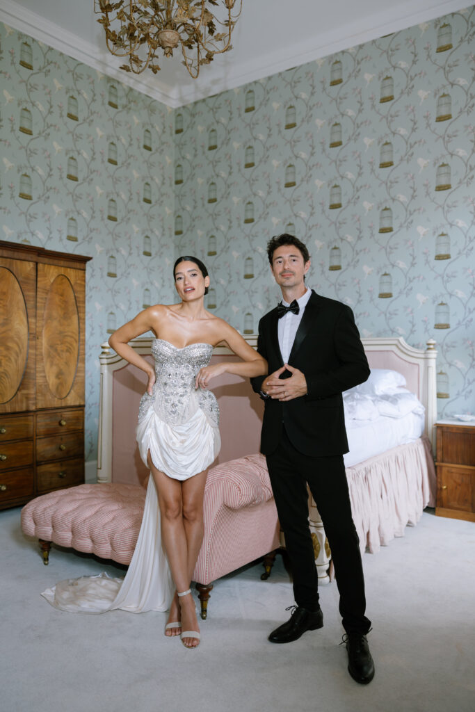 A woman in a strapless, ornate Milla Nova dress and a man in a black tuxedo stand side by side in an elegant, vintage-style bedroom with patterned wallpaper and classic wooden furniture, evoking the charm of a Sandon Hall Wedding.