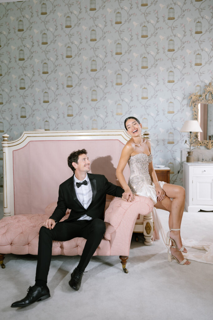 A smiling couple dressed formally sits on a pink upholstered bench in an elegant, vintage-style room. Captured by Rebecca Carpenter Photography, the bride laughs in her strapless Milla Nova dress while holding her partner’s hand.