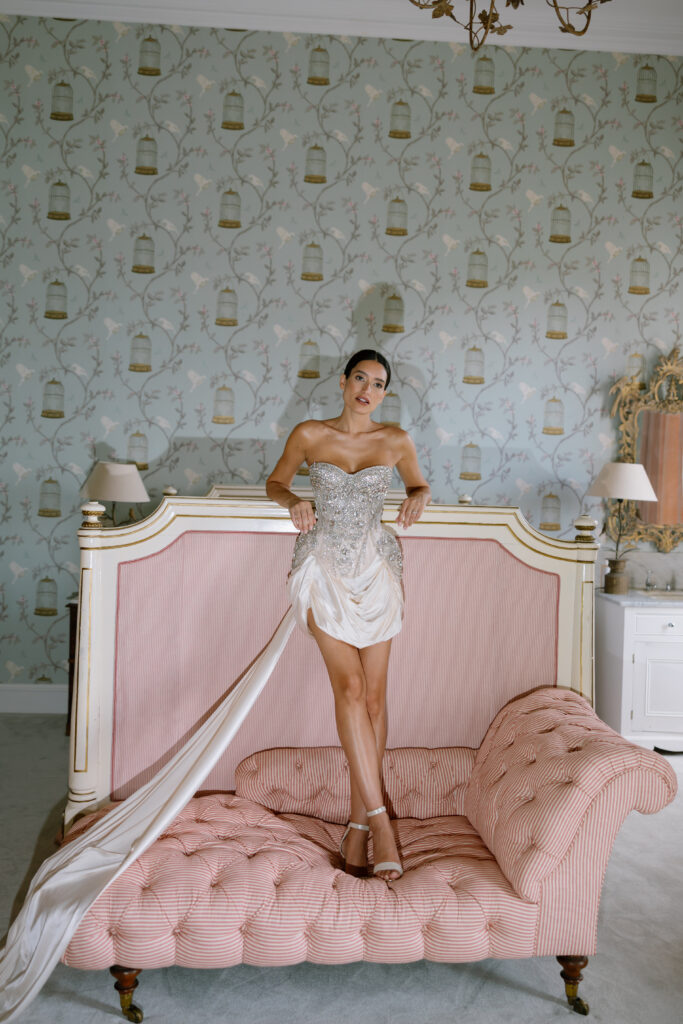 A woman in a strapless, embellished mini dress with a long train stands on a pink tufted chaise lounge in a vintage-style room with birdcage wallpaper, pale furnishings, and elegant decor.